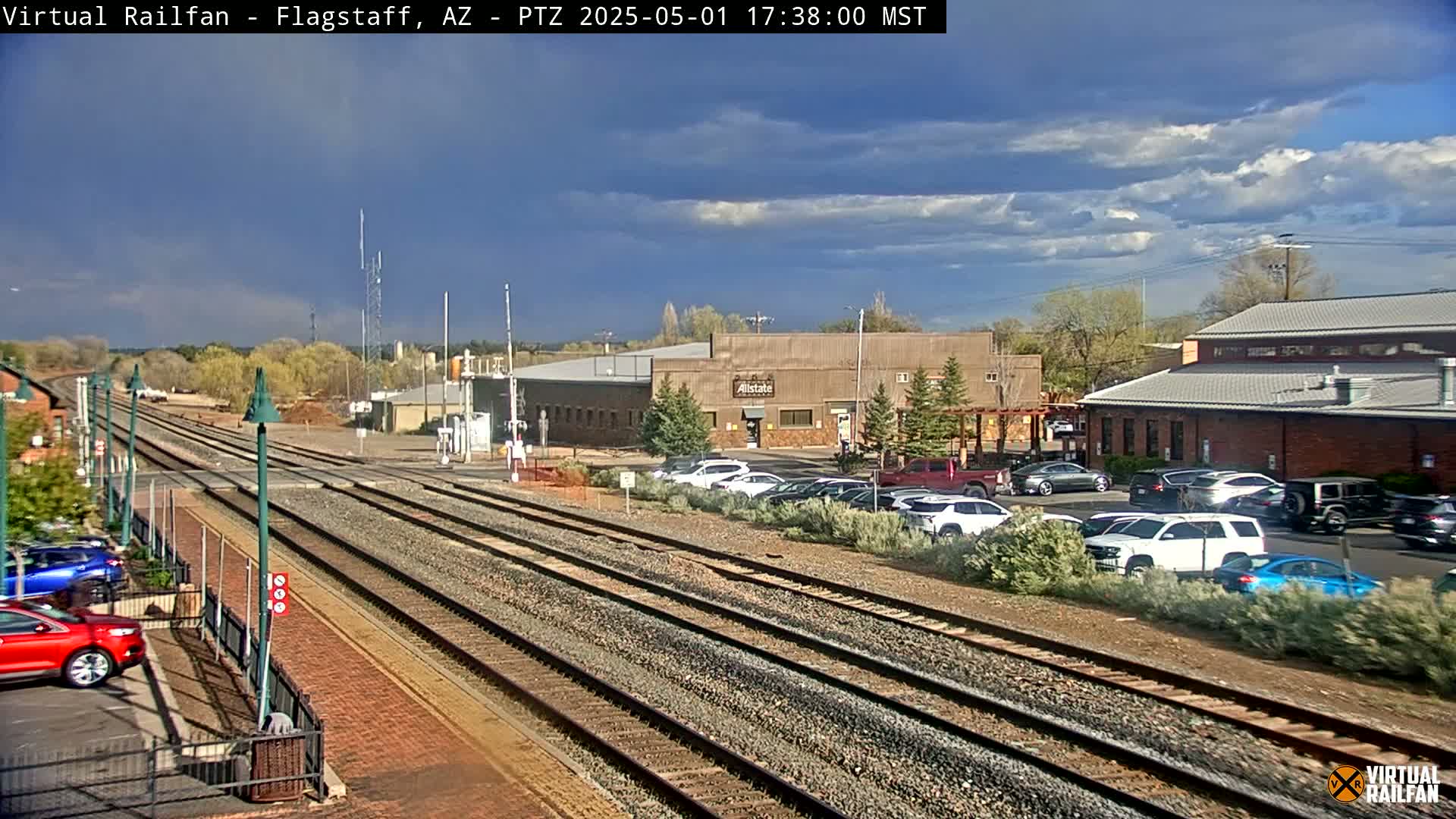 A partly cloudy day shows a railroad track splitting into three paths near several buildings and a parking lot filled with cars.