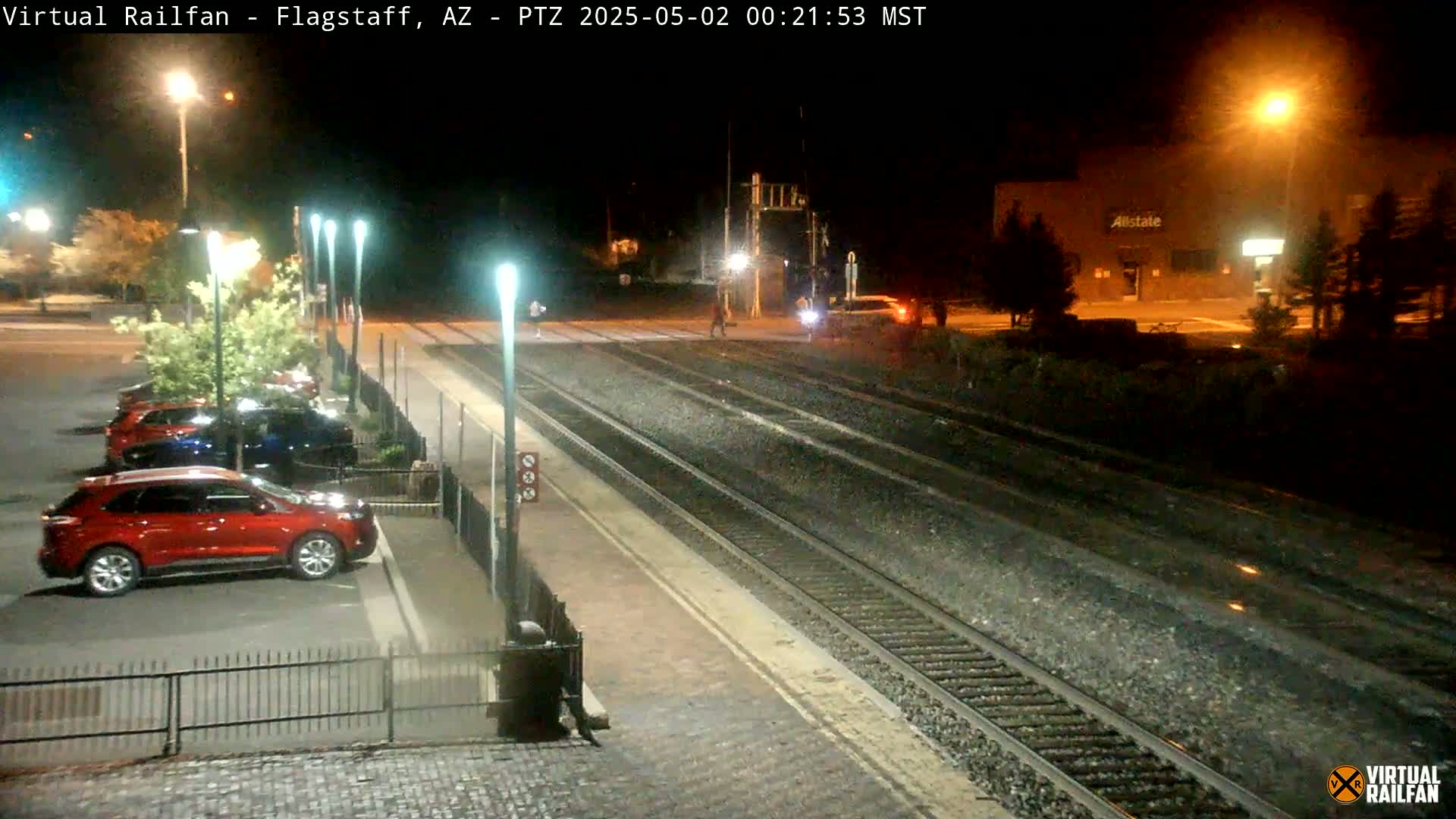 A nighttime view of a train station platform with several cars parked nearby, illuminated by streetlights, under clear skies.