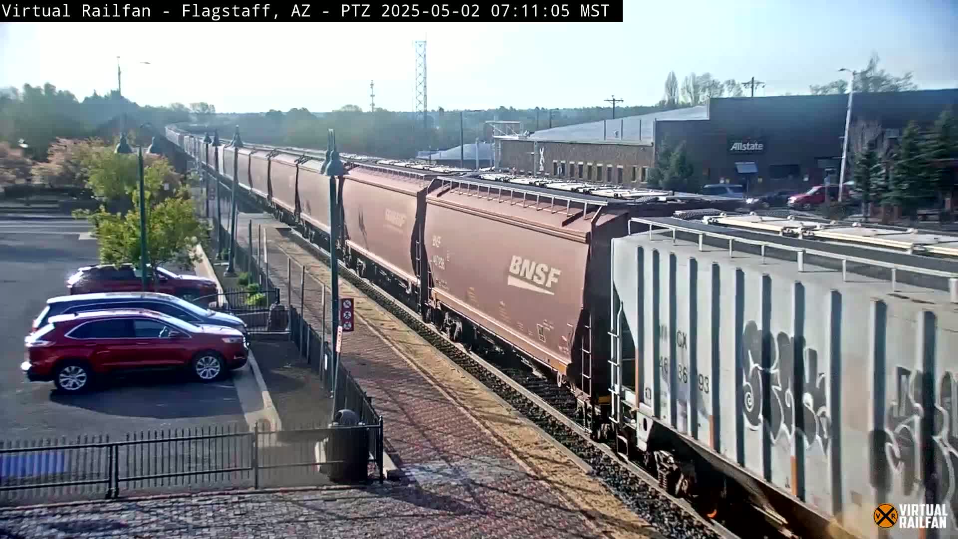 A long freight train passes by a parking lot on a sunny day.