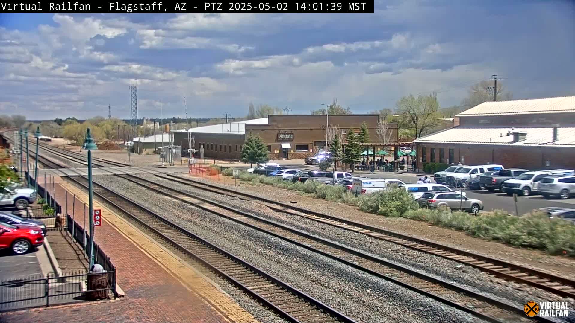 Multiple train tracks run alongside a parking lot filled with cars near several buildings under a partly cloudy sky.