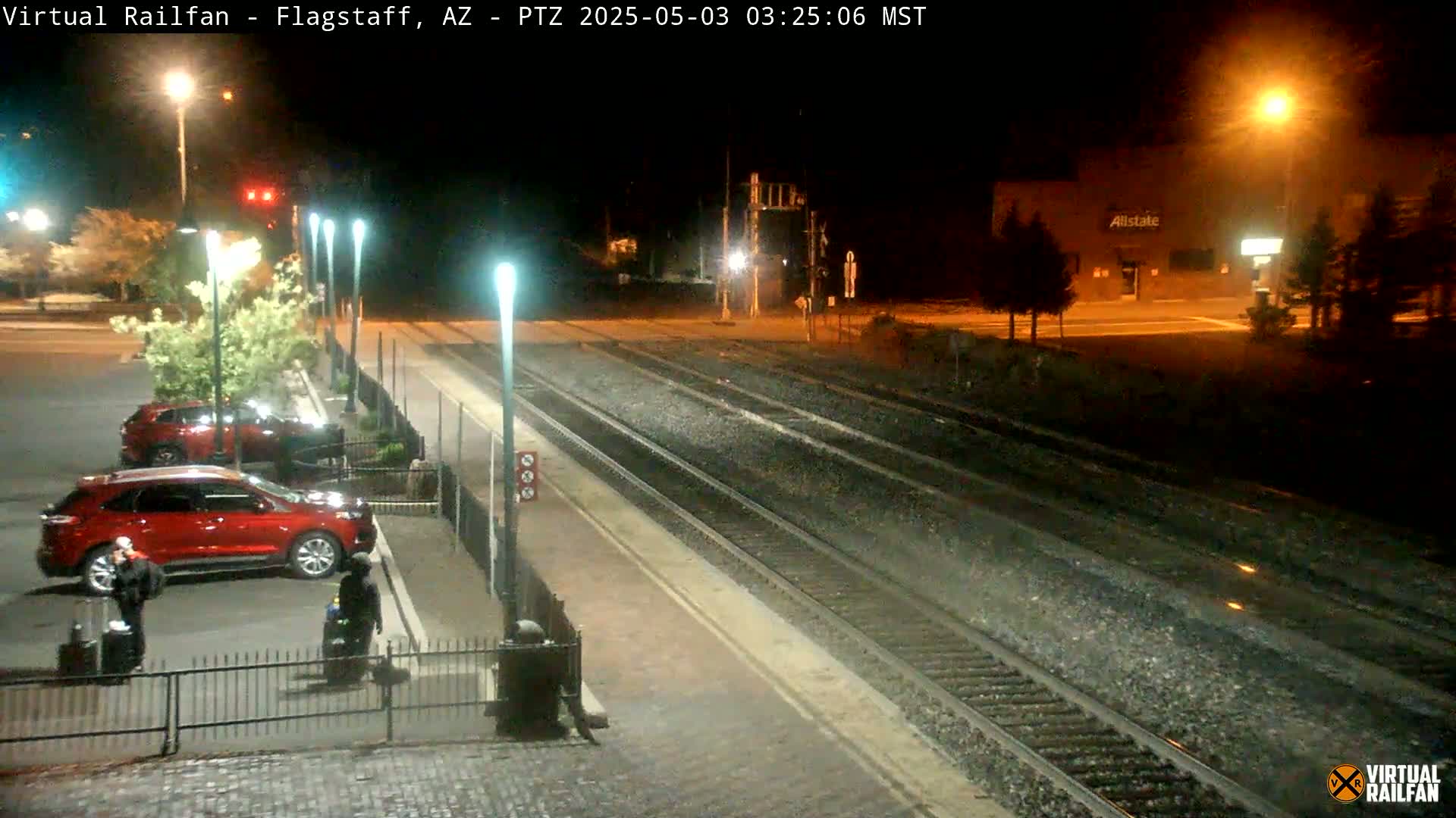 At night, two people with luggage walk past a red SUV parked near train tracks at a station, illuminated by streetlights.