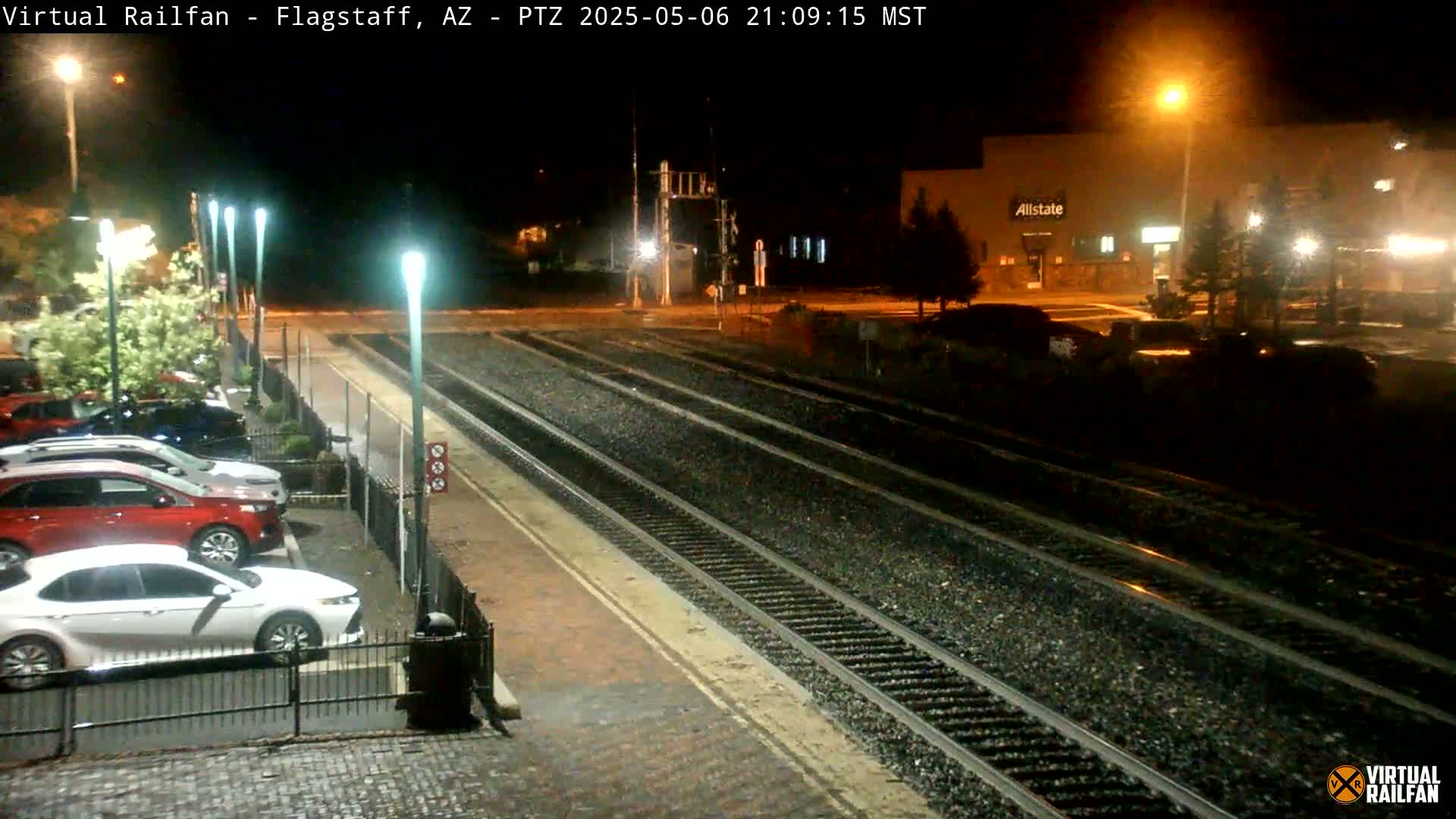 At night, a parking lot with several cars is adjacent to train tracks illuminated by streetlights under a wet, dark sky.