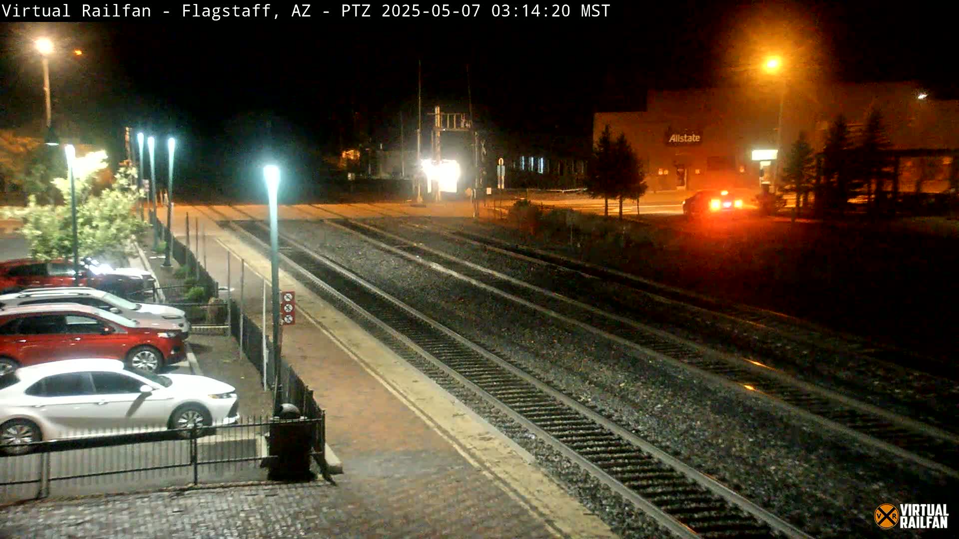 At night, a parking lot next to train tracks is illuminated by streetlights, with several cars parked and a vehicle visible in the distance.