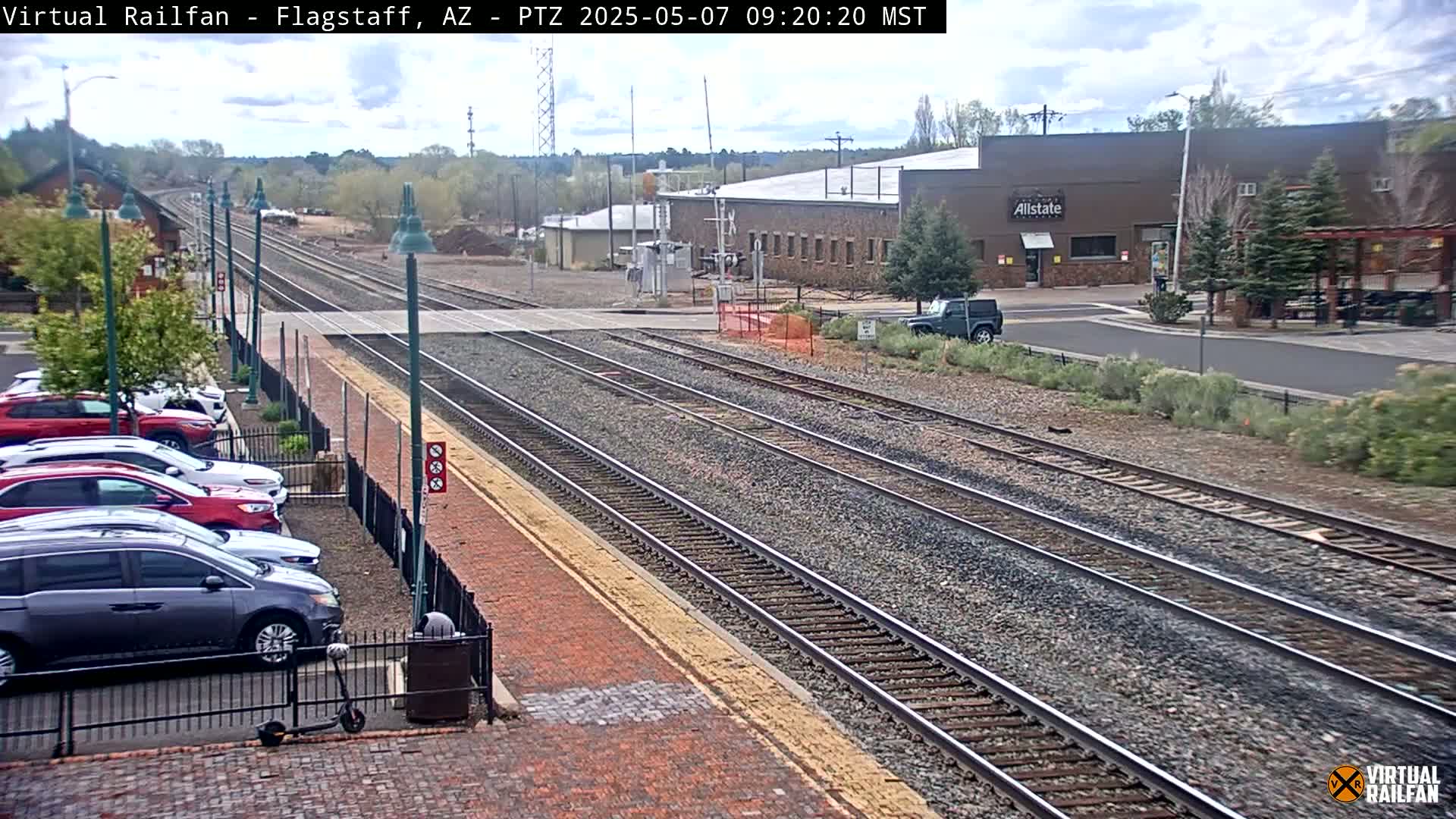 Multiple train tracks extend from a parking lot with several cars parked next to a brick walkway, under a partly cloudy sky.