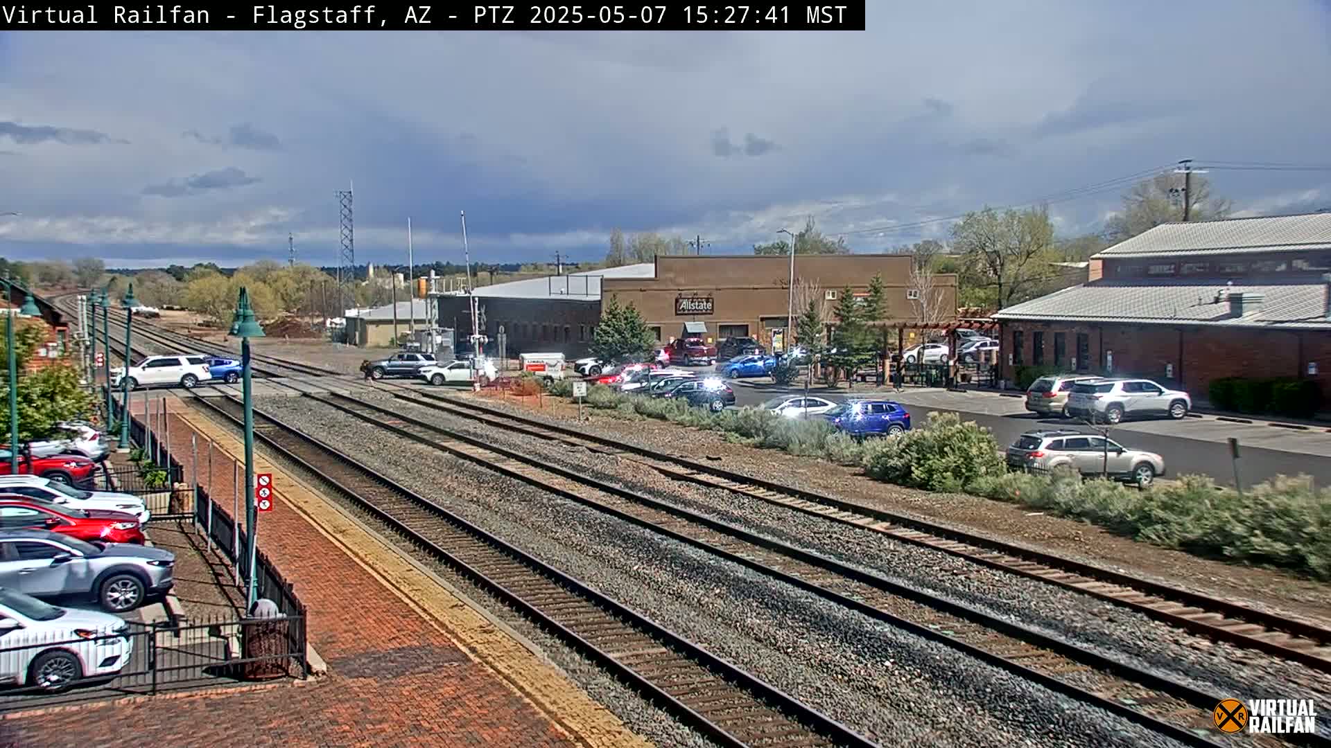 A partly cloudy day reveals a railroad track with multiple train lines, alongside a parking lot filled with cars near several buildings, one of which displays an Allstate logo.