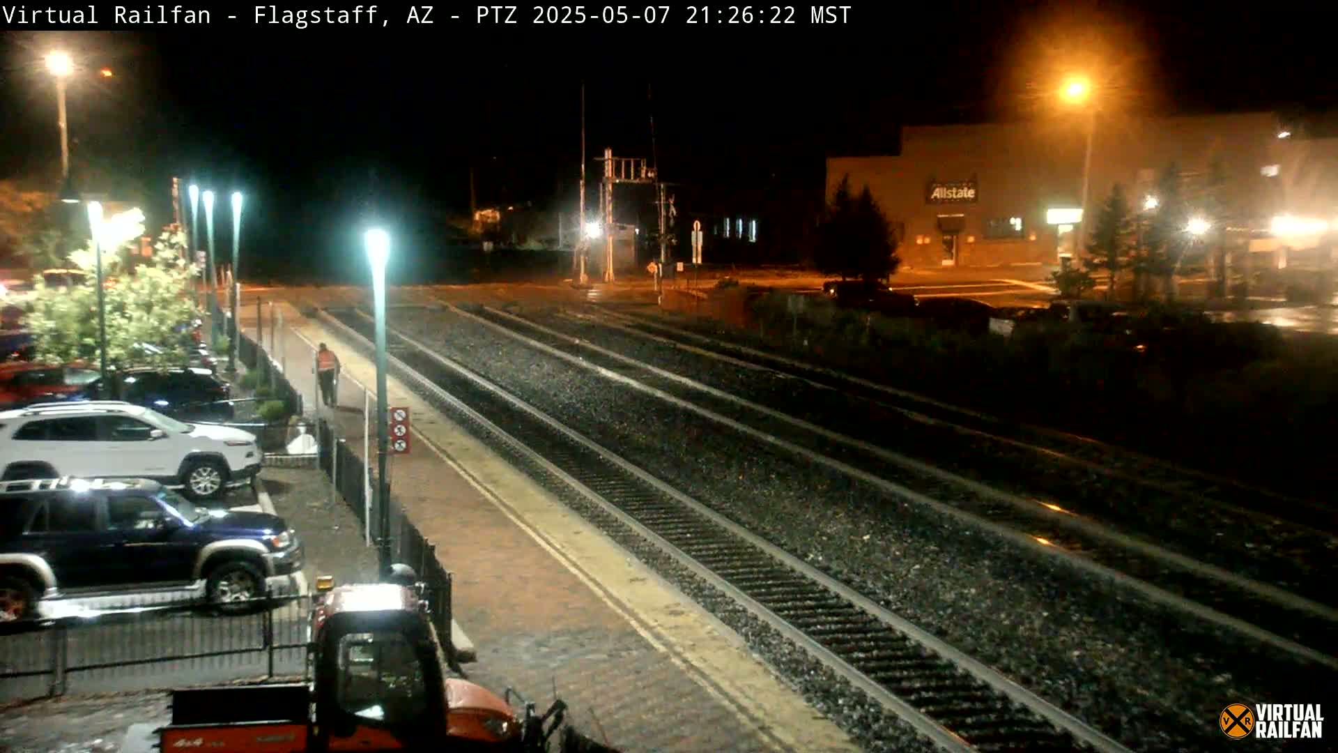 A nighttime view of rain-slicked train tracks, a parking lot with several vehicles, and a person walking along a platform under illuminated streetlights.