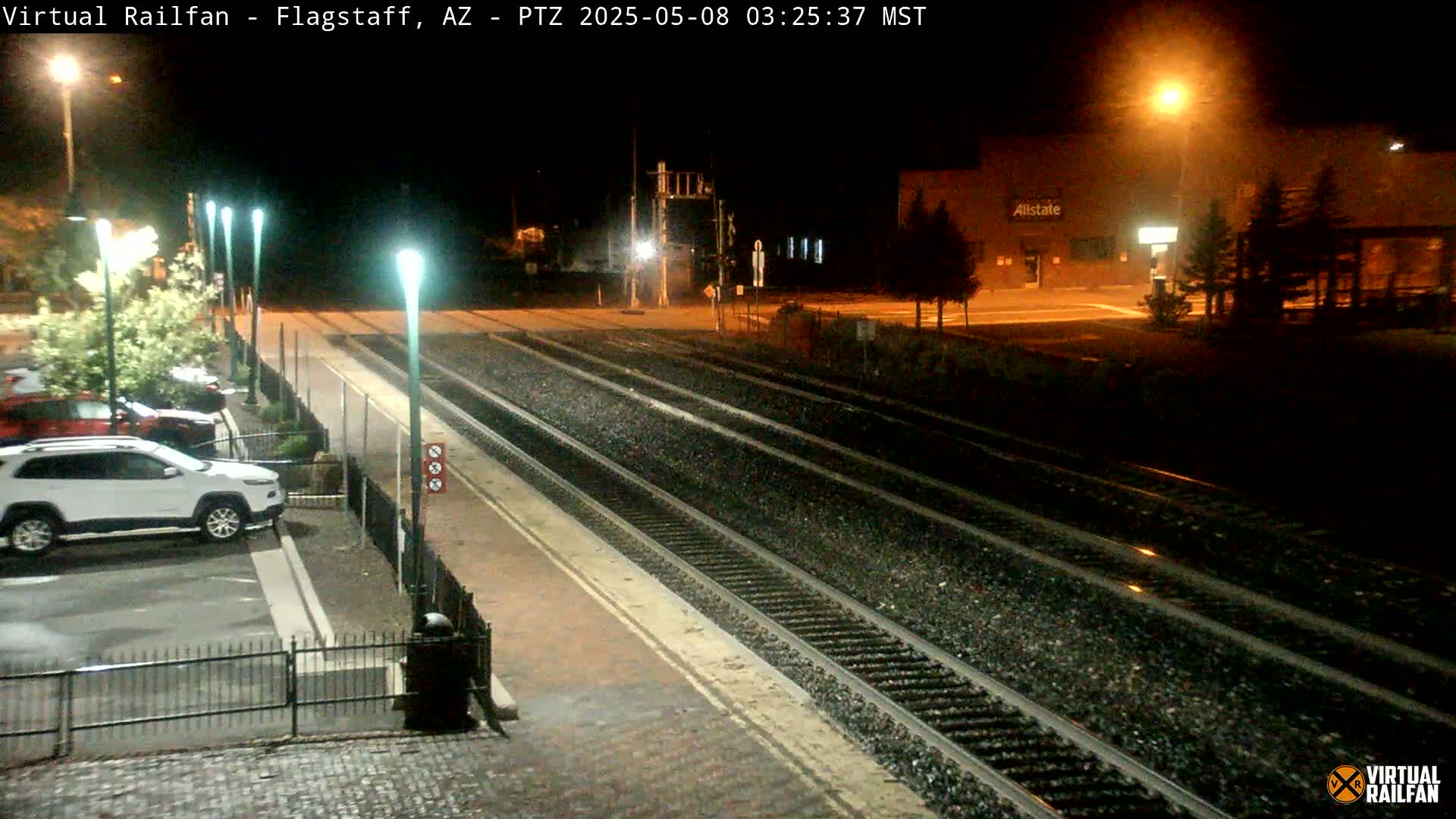 At night, a train station platform with several cars parked nearby is illuminated by streetlights, with multiple train tracks extending into the distance.