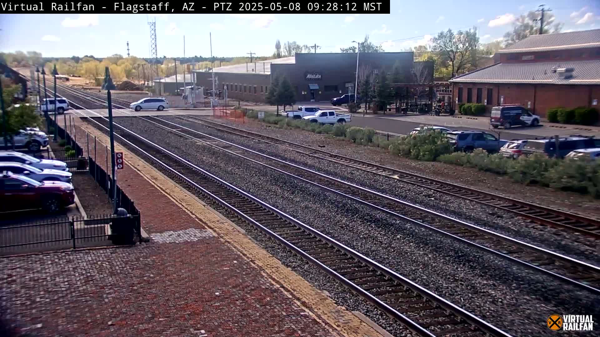Multiple train tracks run through a parking lot next to a brick building and several other commercial structures under a mostly sunny sky.