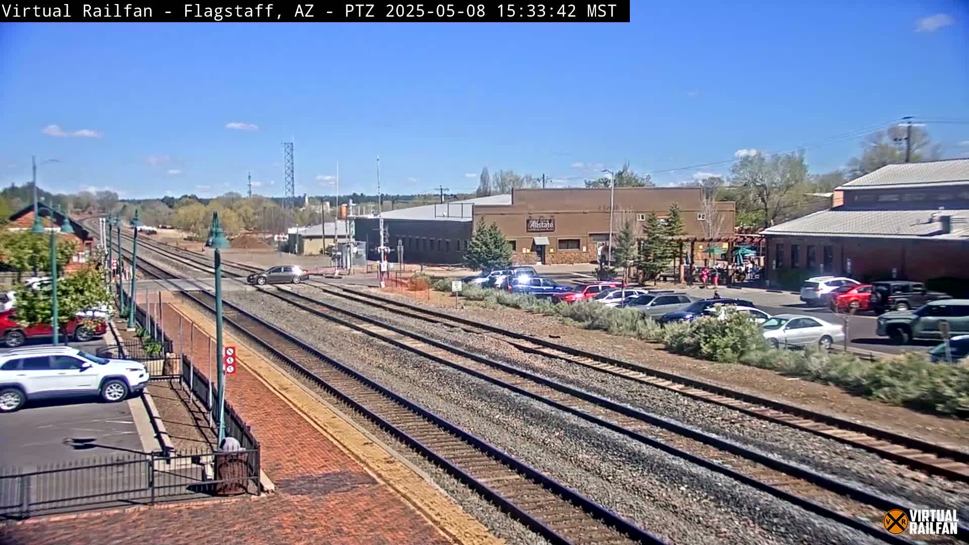A sunny day reveals multiple train tracks alongside a road with parked cars and buildings, including one with a visible business sign.