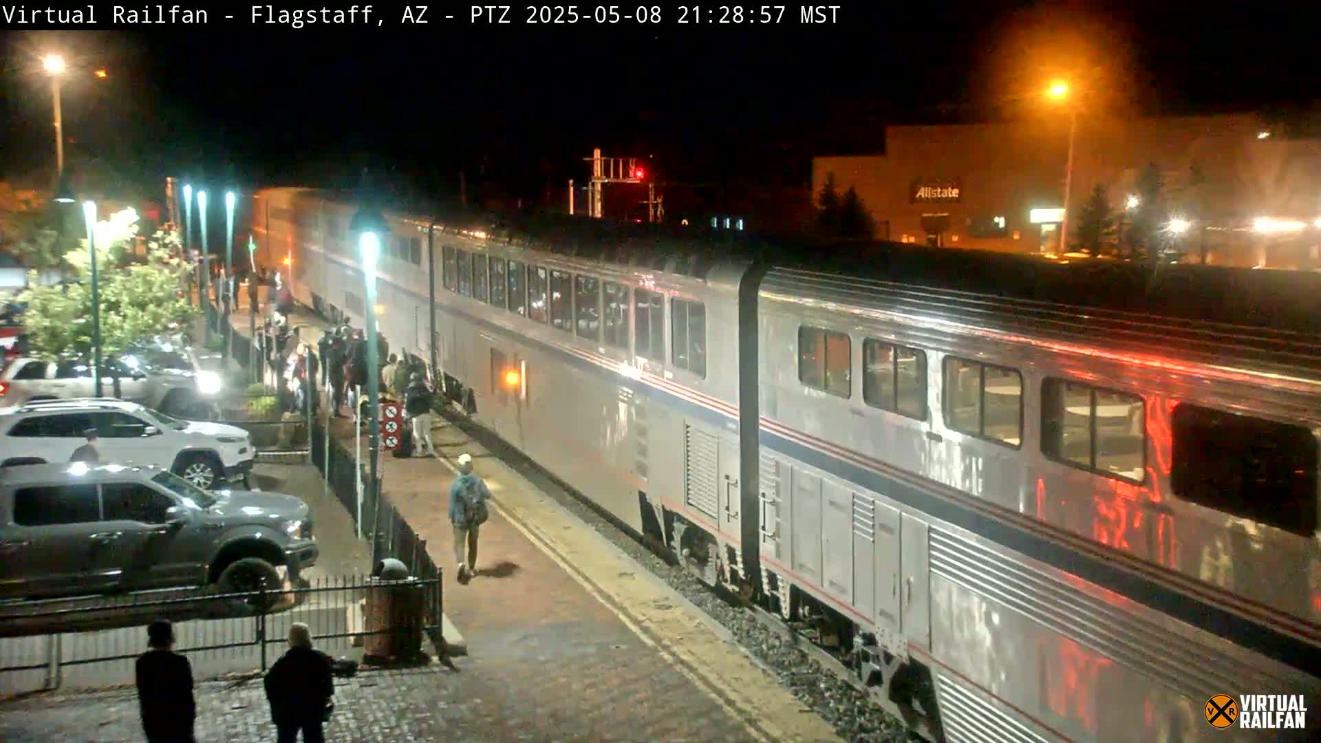 A long, silver passenger train is at a station at night, with people standing on the platform and several vehicles parked nearby.