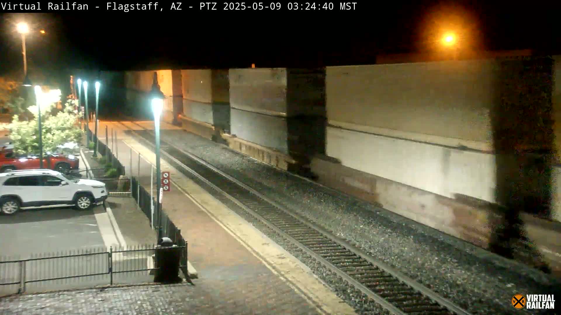 A nighttime view of a train passing by a station platform with several cars parked nearby.