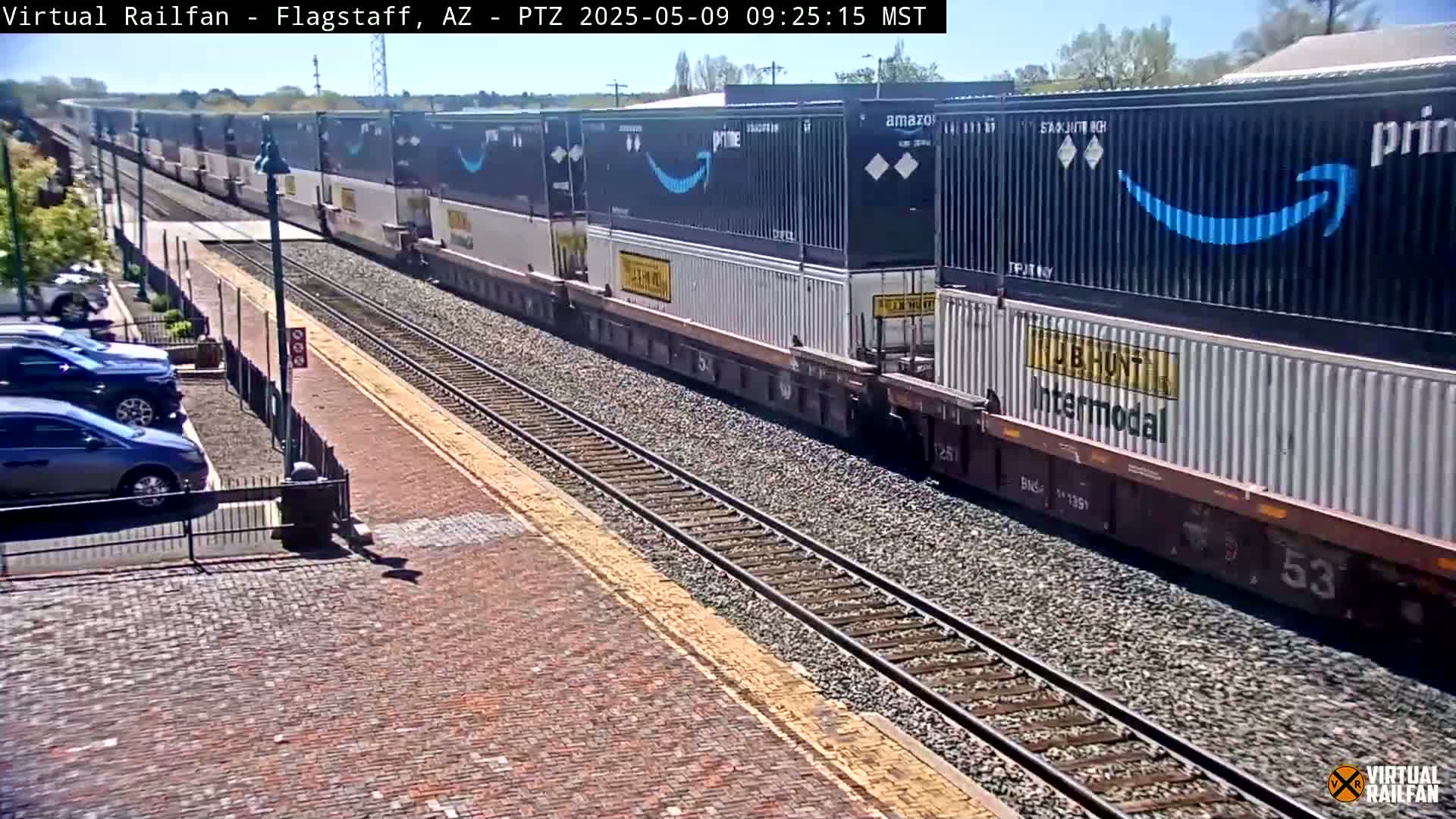 A long freight train carrying numerous shipping containers, some of which are Amazon Prime branded, passes by a brick paved area with parked cars under a sunny sky.