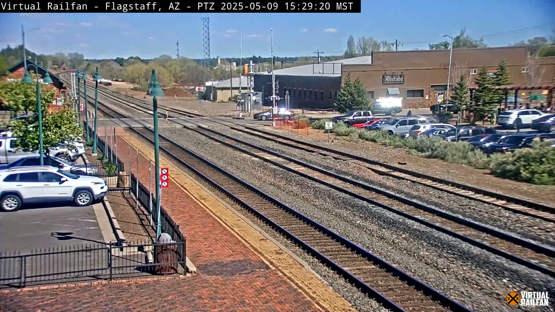 A sunny day reveals multiple train tracks extending into the distance, alongside a parking lot with cars and a brick roadway, all situated near some buildings.