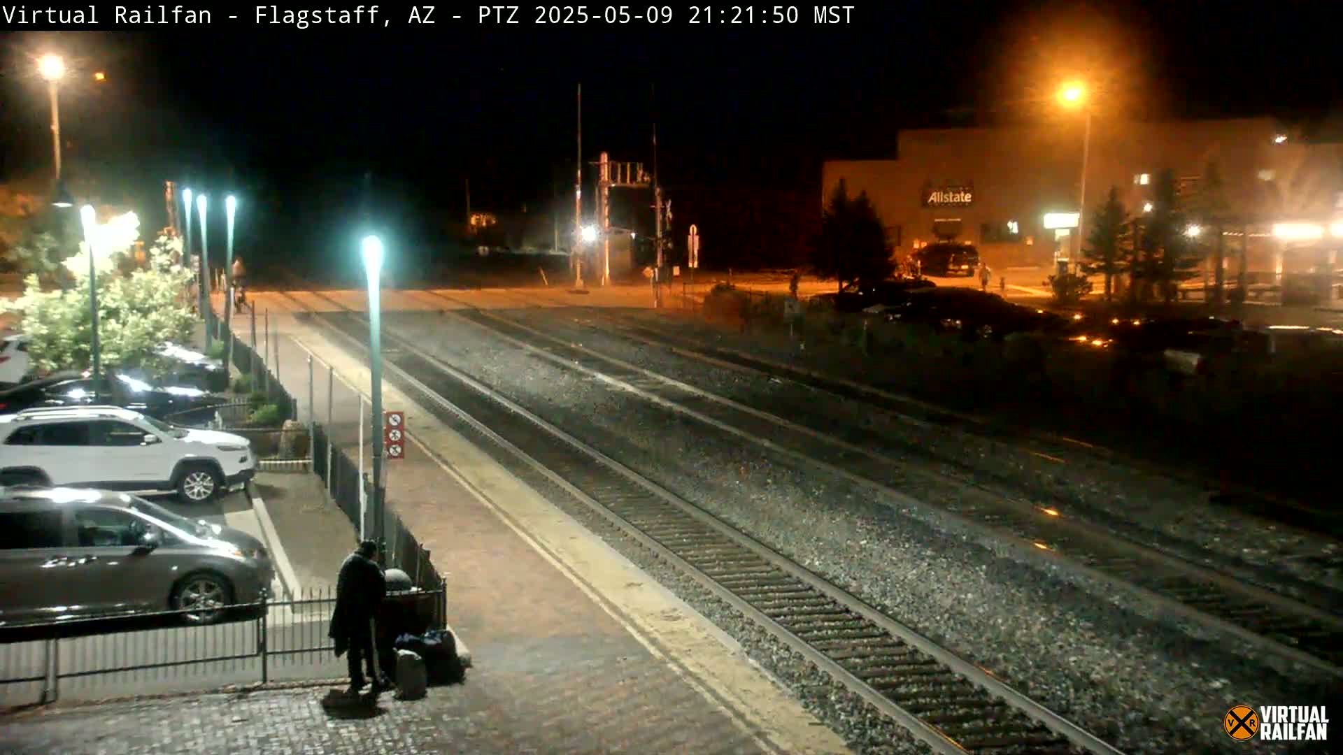 A nighttime view of a train station with several cars parked nearby and a person standing with luggage near a fence beside railroad tracks.
