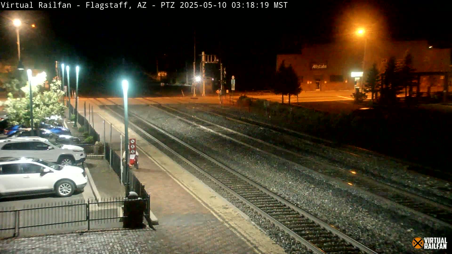 A nighttime view of a train station with several cars parked nearby and train tracks extending into the distance under artificial light.
