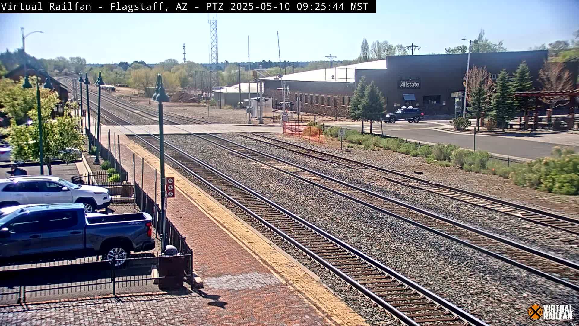 A sunny day view of multiple train tracks next to a parking lot with cars, and a building in the background.