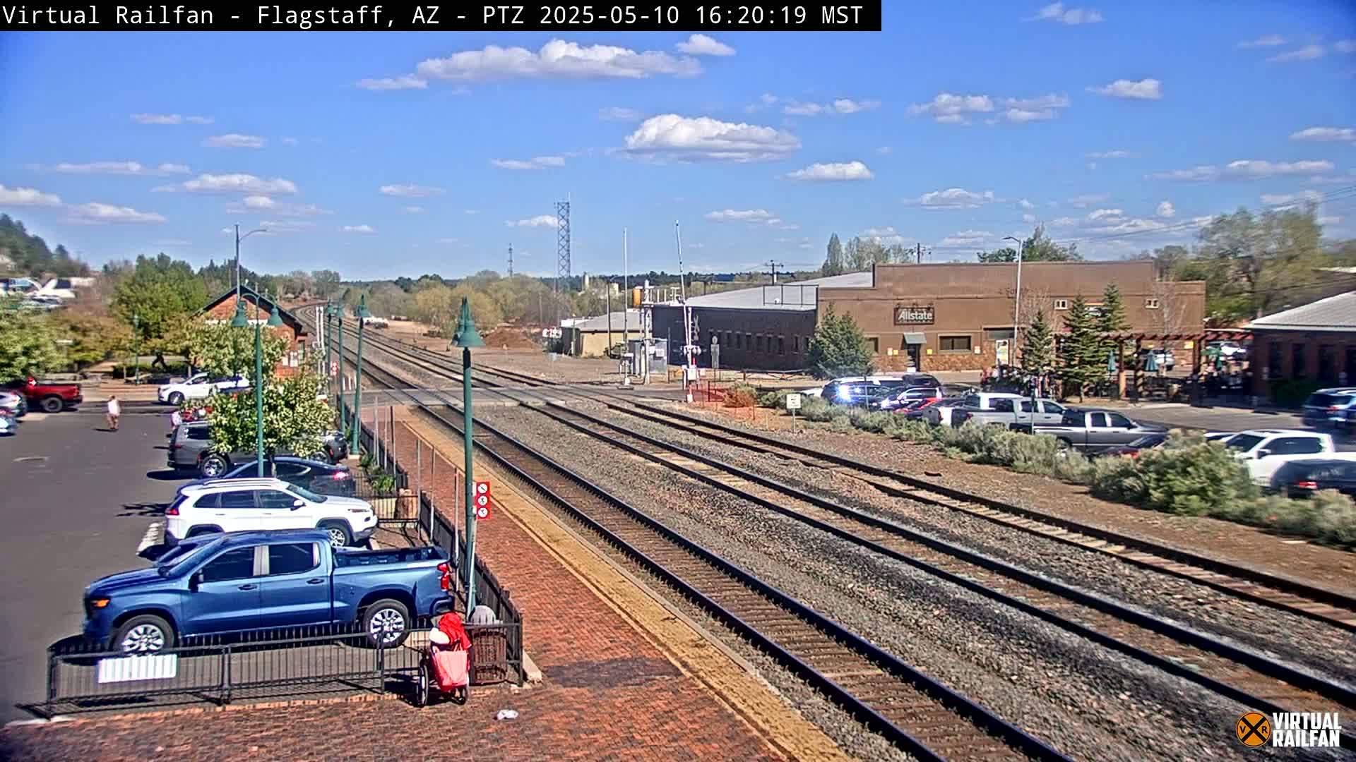 A sunny day at a train station with multiple train tracks, parked cars, and a few pedestrians.