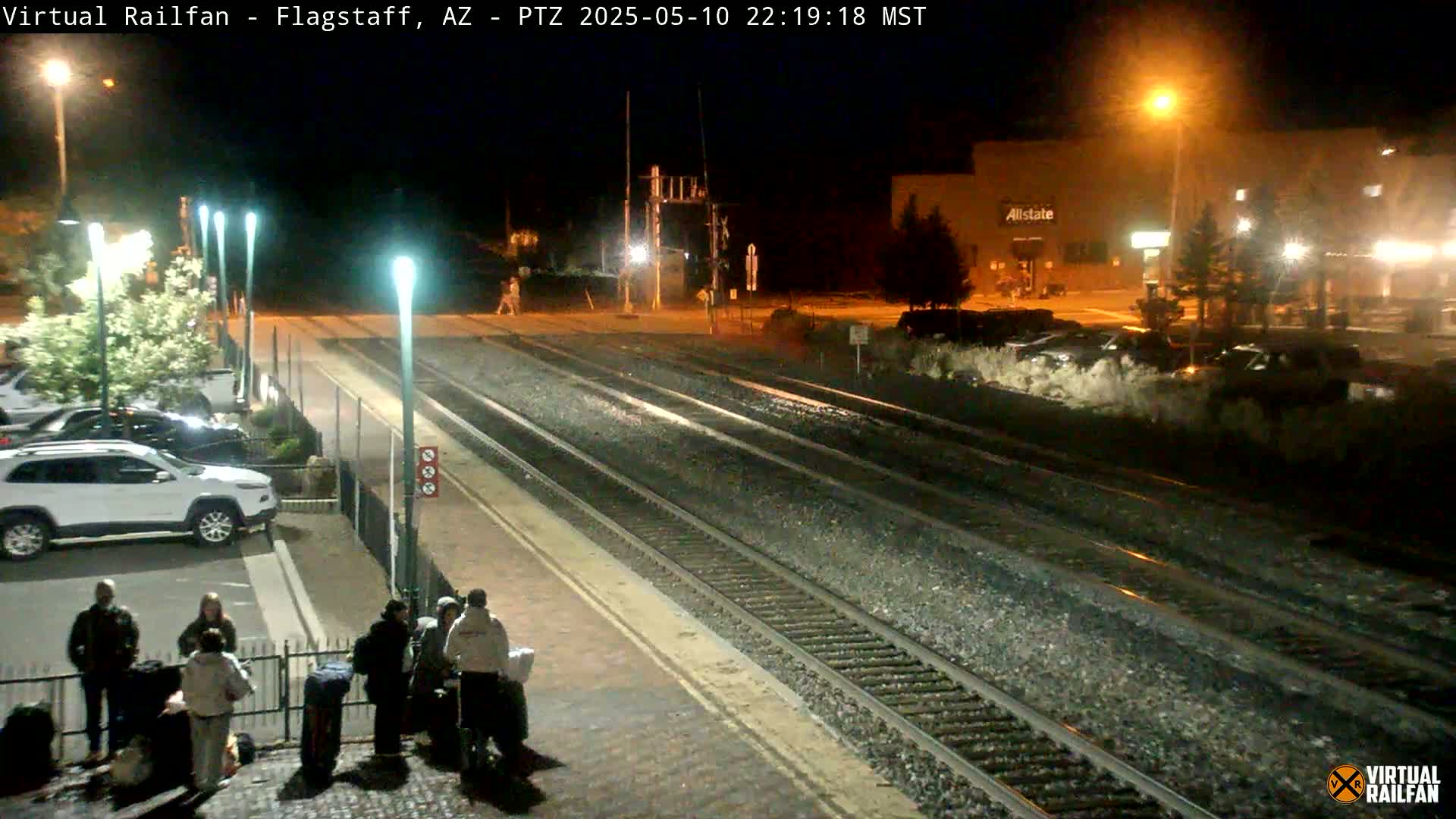 A nighttime view of a train station platform with several people standing near luggage, illuminated by streetlights, with train tracks extending into the distance.