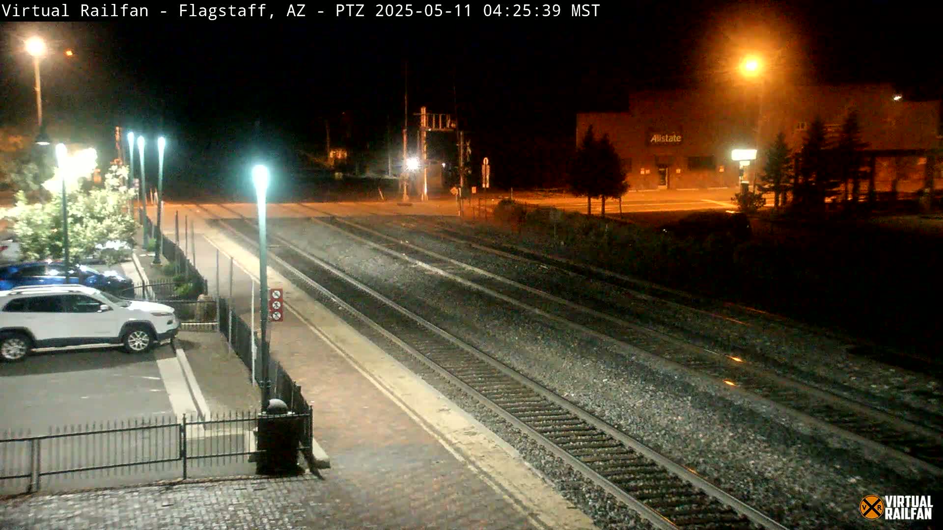 A nighttime view of train tracks at a station with cars parked nearby, illuminated by streetlights.