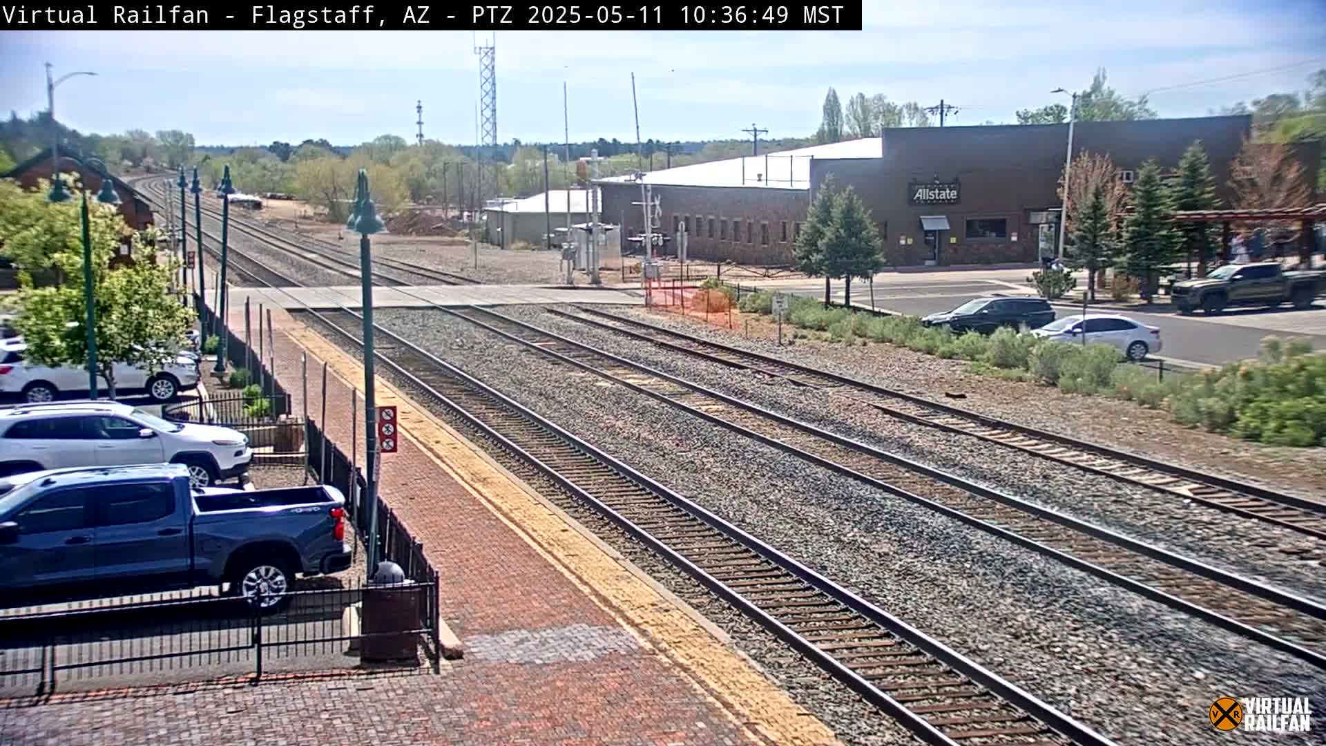 A sunny view of several train tracks next to a parking lot with cars and a building, all surrounded by low vegetation.