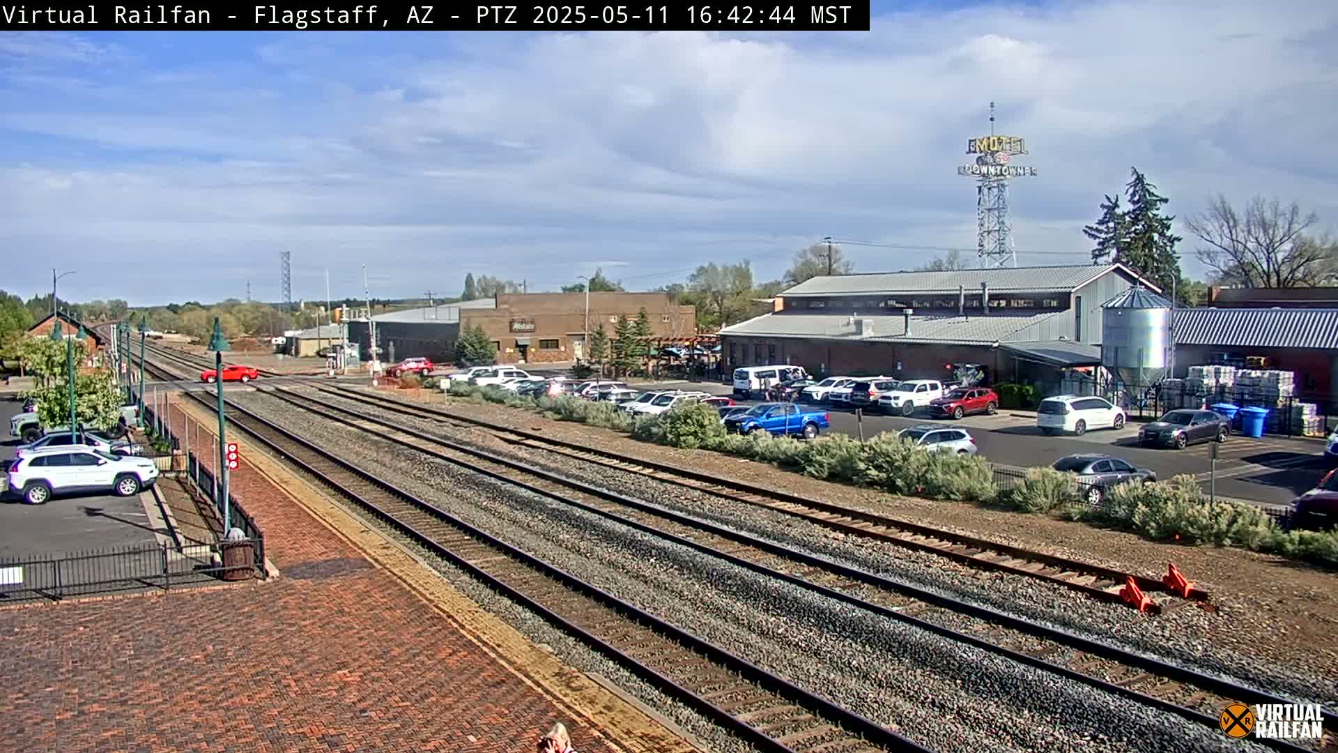 A sunny day reveals railroad tracks intersecting with a street, alongside a parking lot filled with cars and several buildings, including one with a silo.