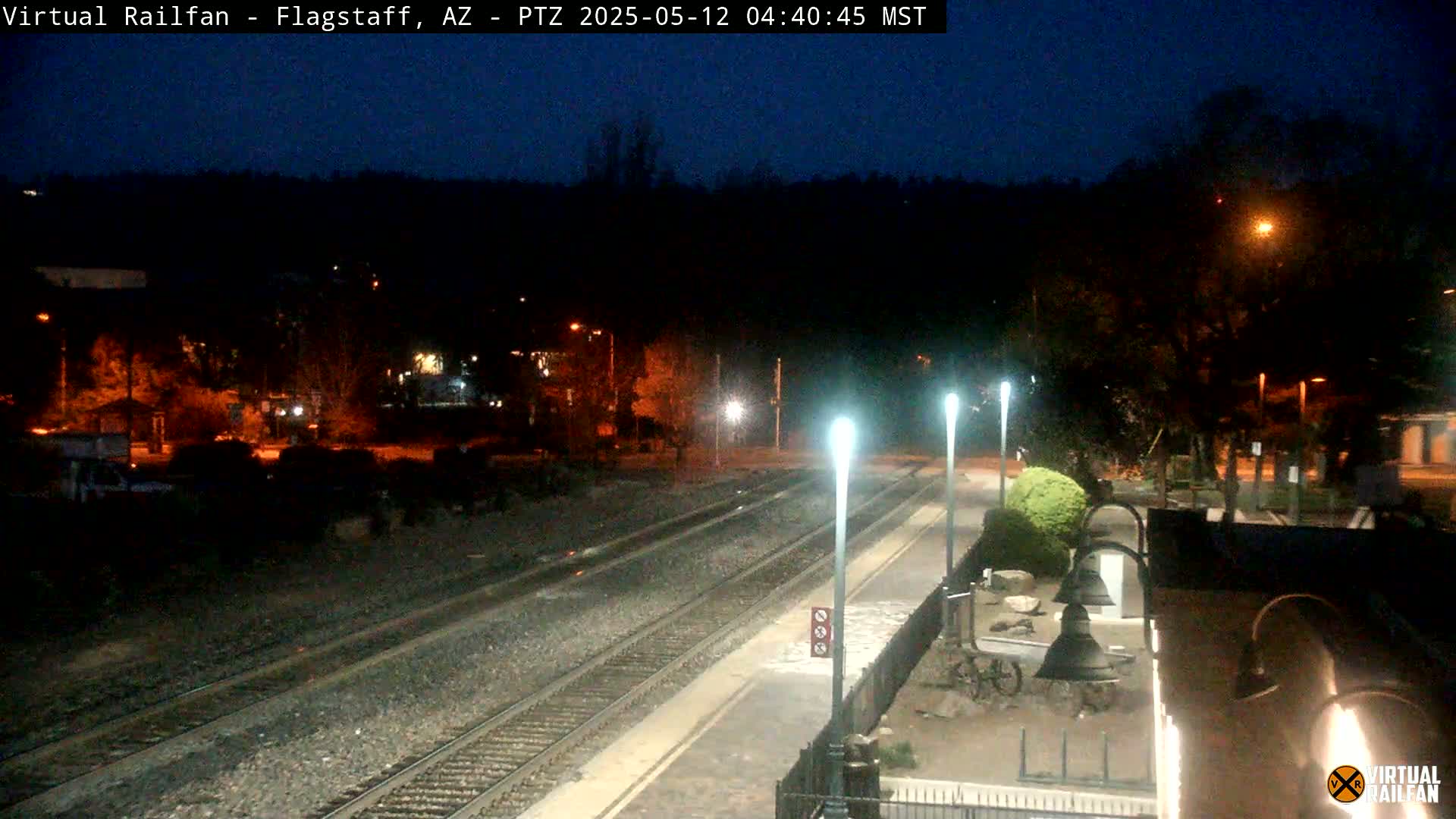 A nighttime view of a train station platform with two sets of train tracks, illuminated by streetlights against a dark, tree-lined background.