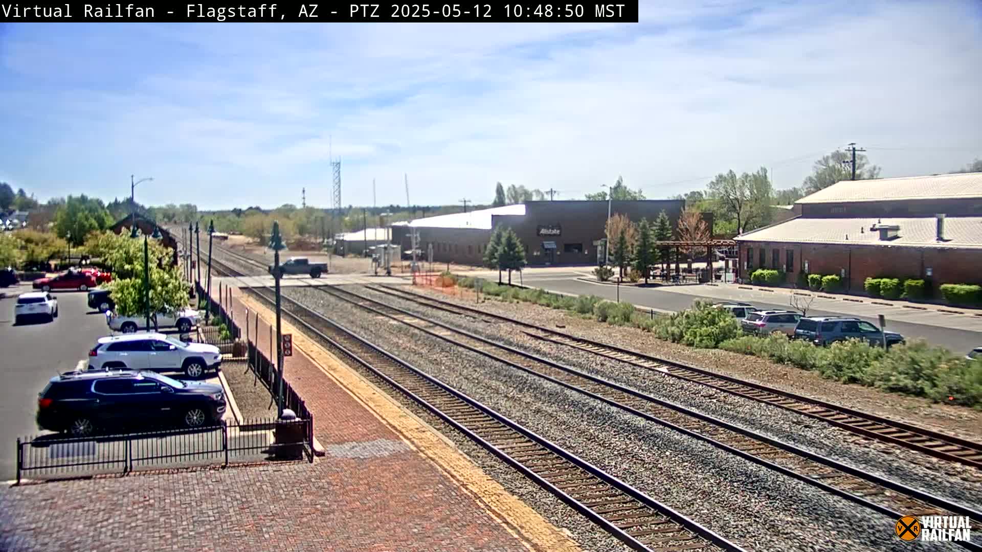 A sunny day shows train tracks that extend into the distance past a parking lot with several cars, and various buildings including what looks like a warehouse and train depot.