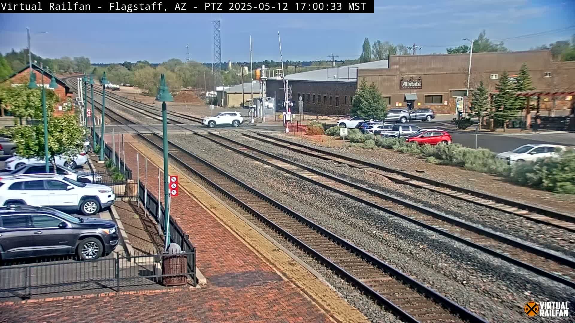 A sunny day reveals multiple train tracks intersecting a roadway, with parked cars lining a brick street next to a railway station.