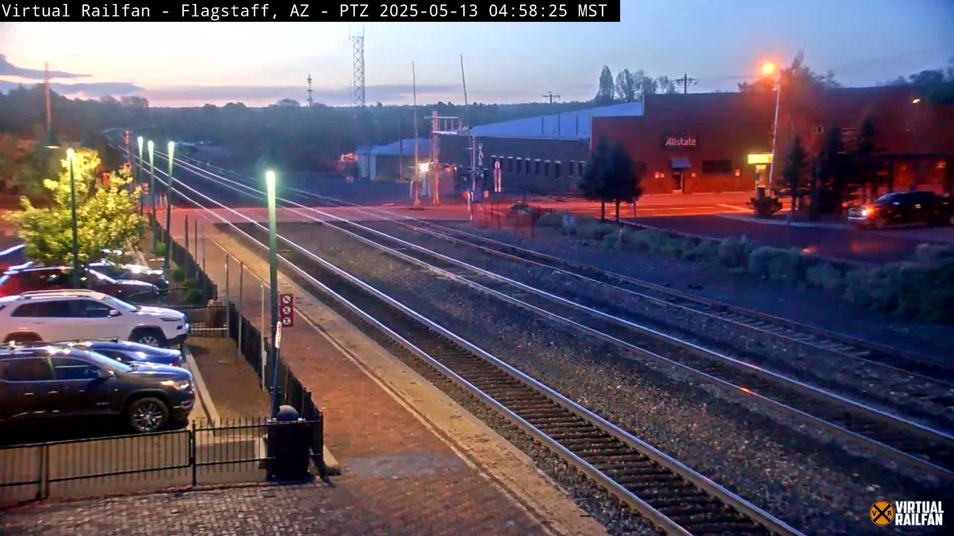 Several cars are parked in a lot next to train tracks at dusk.