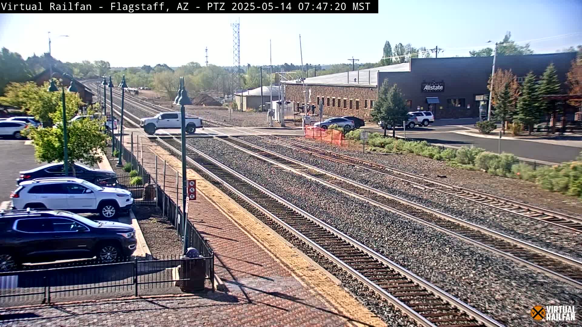 Multiple sets of train tracks run through a parking lot with several vehicles parked nearby under a sunny sky.