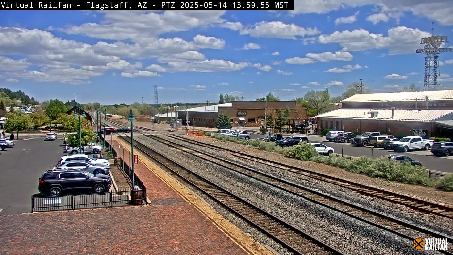 A sunny day with partly cloudy skies shows a railroad track extending into the distance, past a parking lot with cars, and various buildings, including a large brick structure.