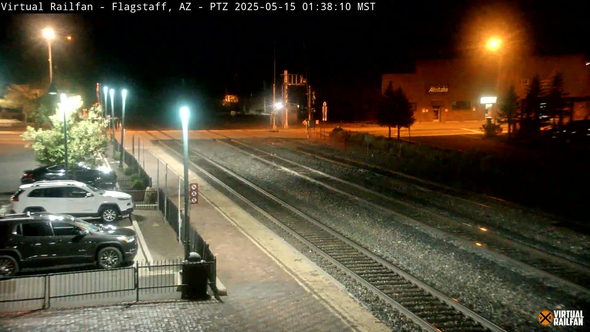 A nighttime view of a train station platform with several cars parked nearby, under bright artificial lights.