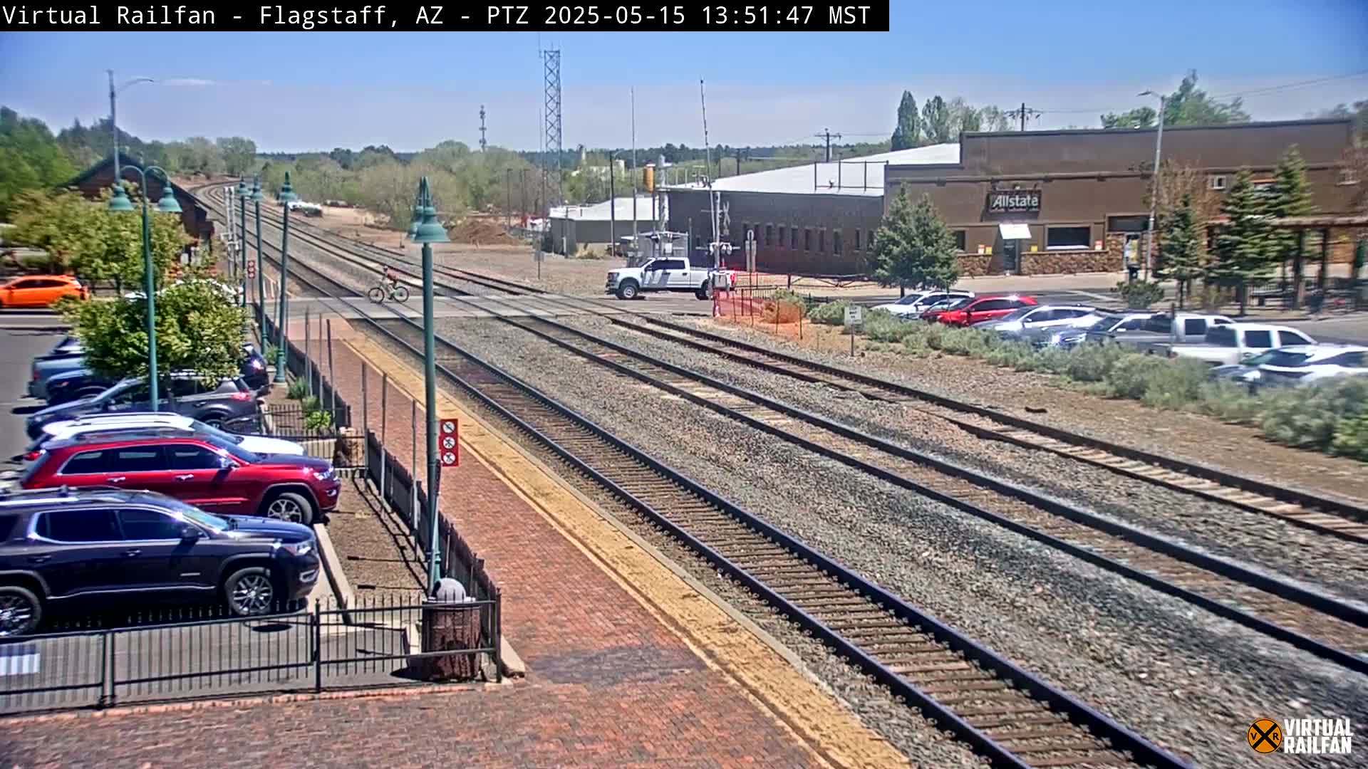 A sunny day reveals multiple train tracks intersecting a roadway with cars parked nearby and a cyclist crossing the tracks.