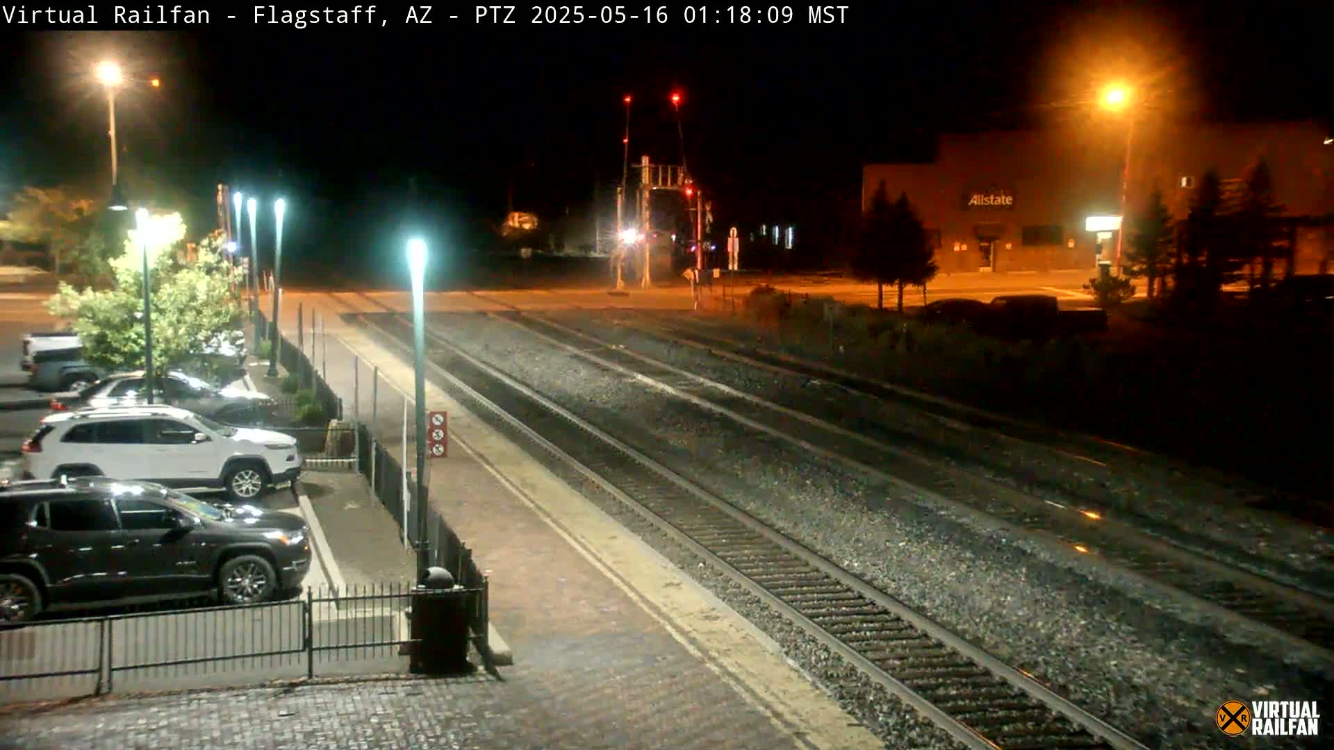 A nighttime view of a train station platform with several parked cars, train tracks, and streetlights illuminating the scene.