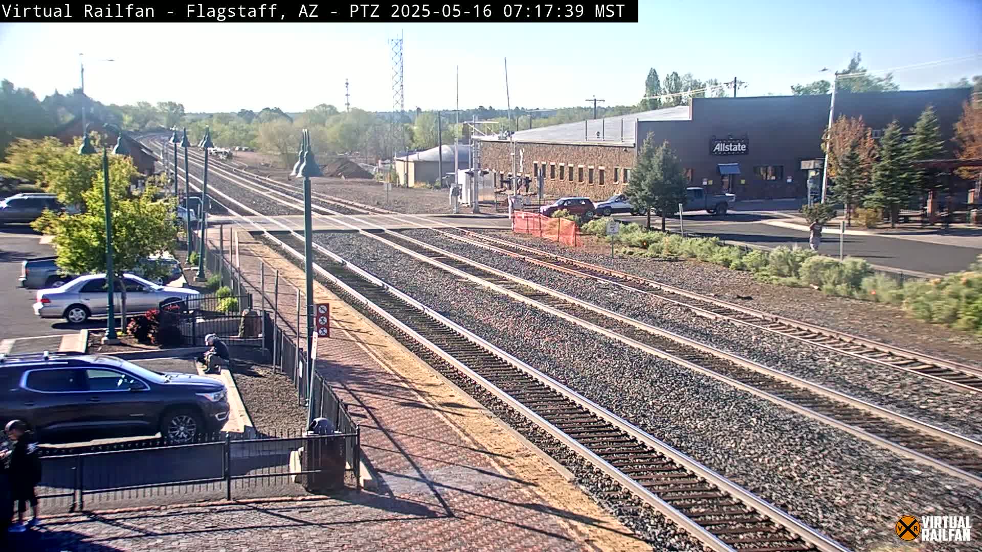 A sunny outdoor view of multiple train tracks leading to a station, with cars parked nearby and a few buildings visible in the distance.