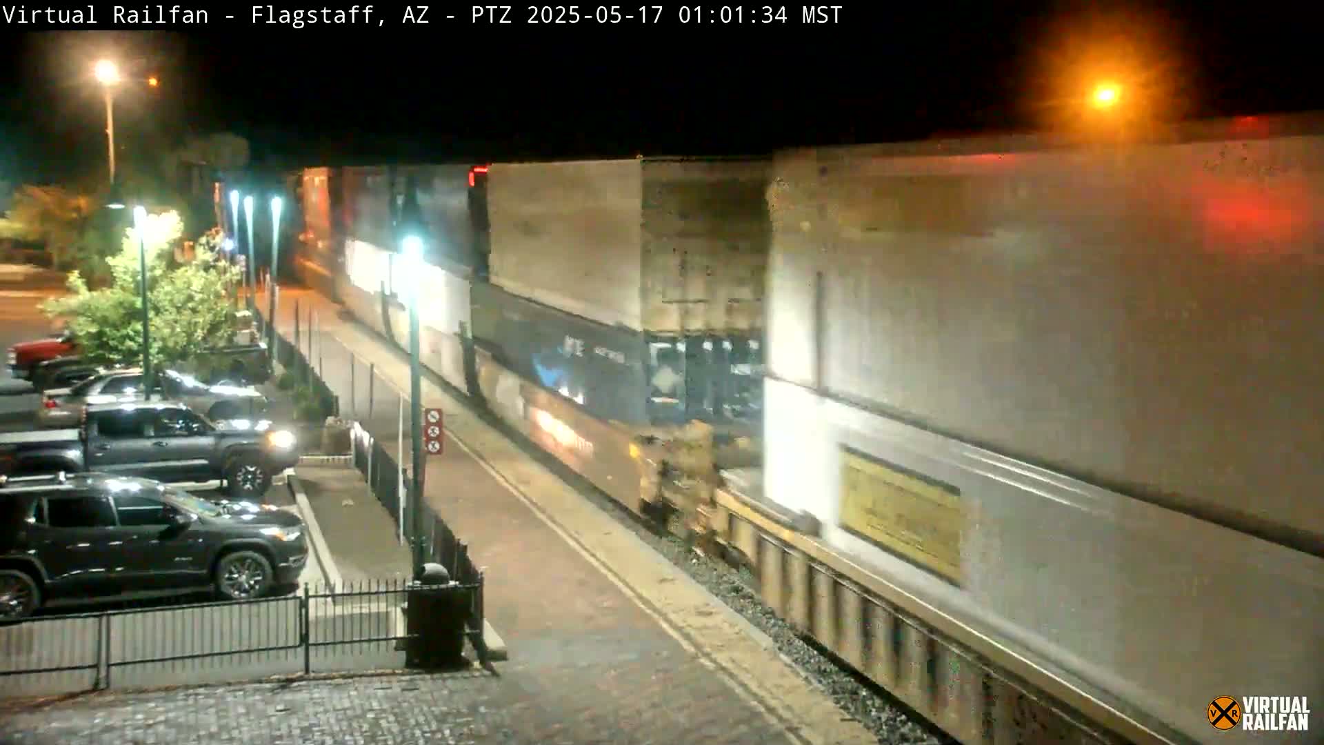 At night, a long freight train passes by a paved area with parked cars next to a building under bright streetlights.