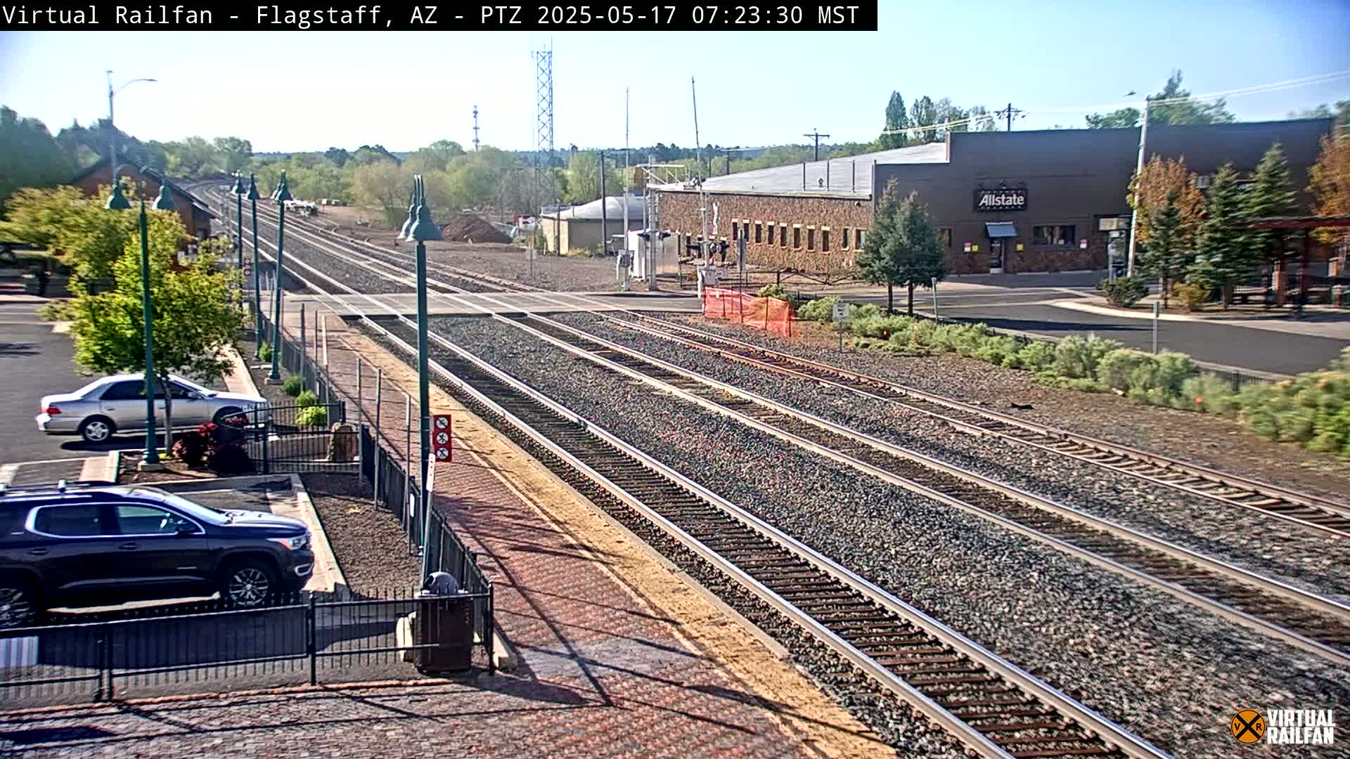 Multiple train tracks extend into the distance on a sunny day, with cars parked nearby and low buildings visible in the background.