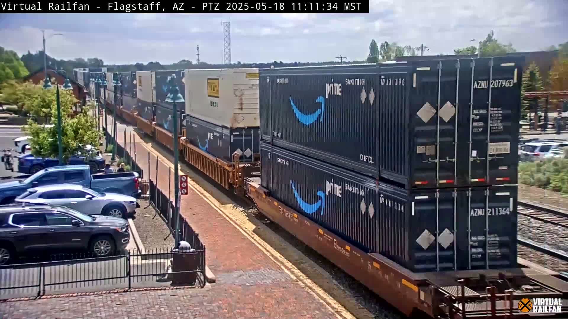 A freight train carrying stacked shipping containers, some bearing the Amazon Prime logo, passes along a brick roadway next to a parking lot on a partly sunny day.