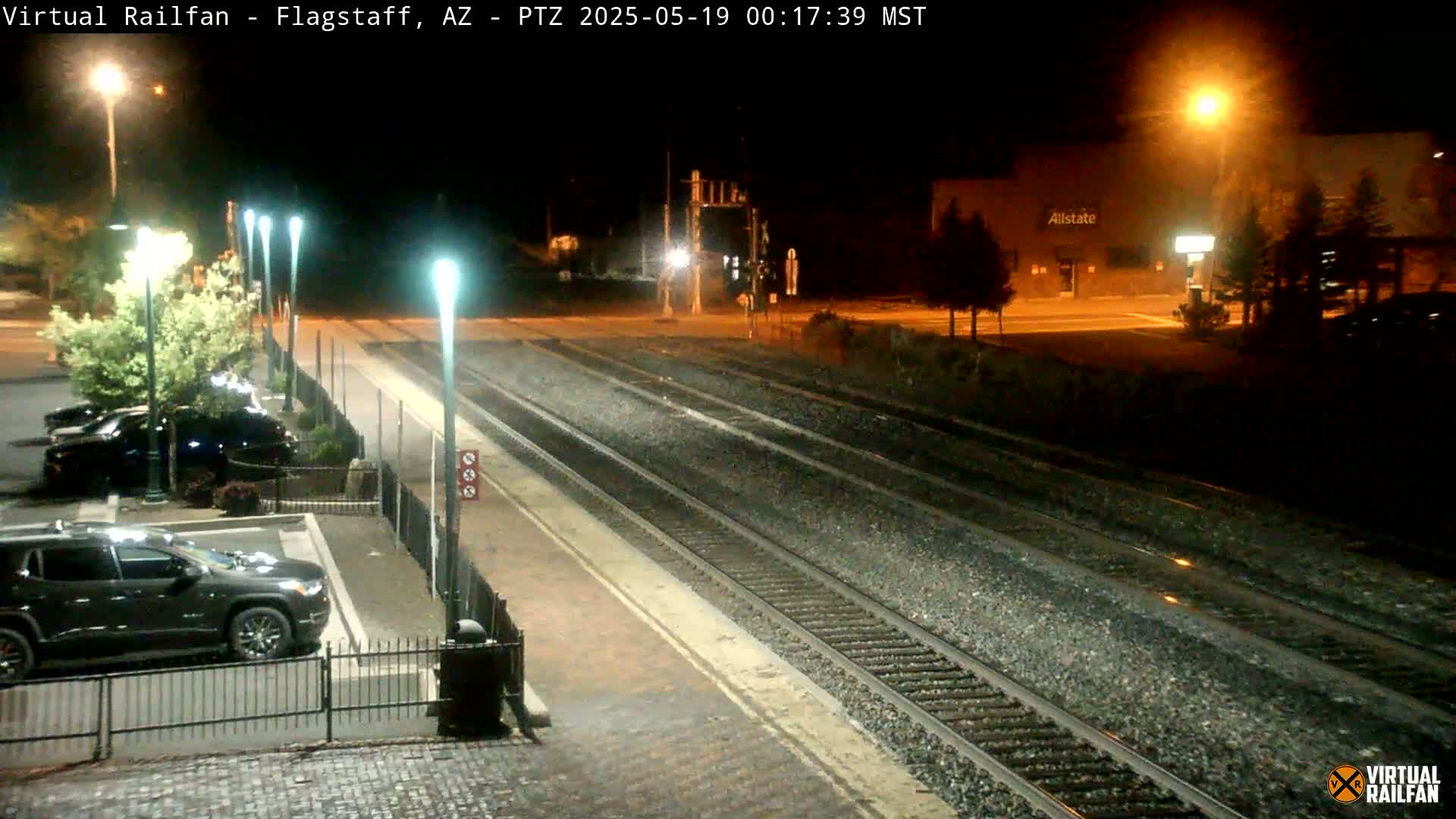 A nighttime view of a train station platform with multiple tracks, several parked cars, and streetlights illuminating the scene.