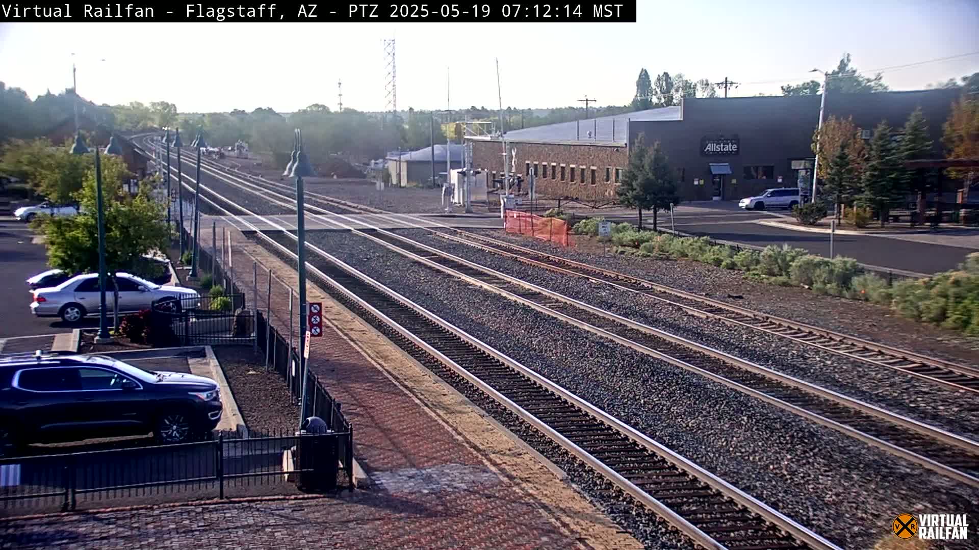 Multiple train tracks extend into the distance under a partly sunny sky, with several cars parked in a lot next to a building.