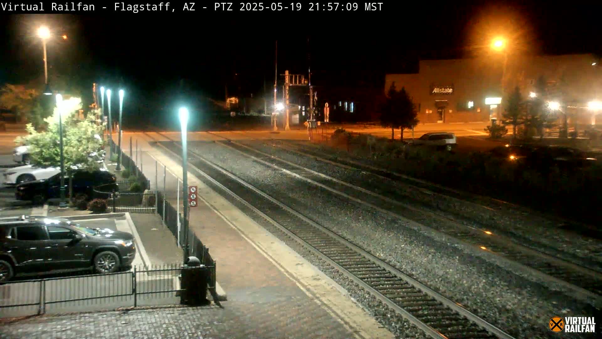 A nighttime view of a train station with multiple sets of train tracks, parking lot with cars, and buildings illuminated by streetlights.