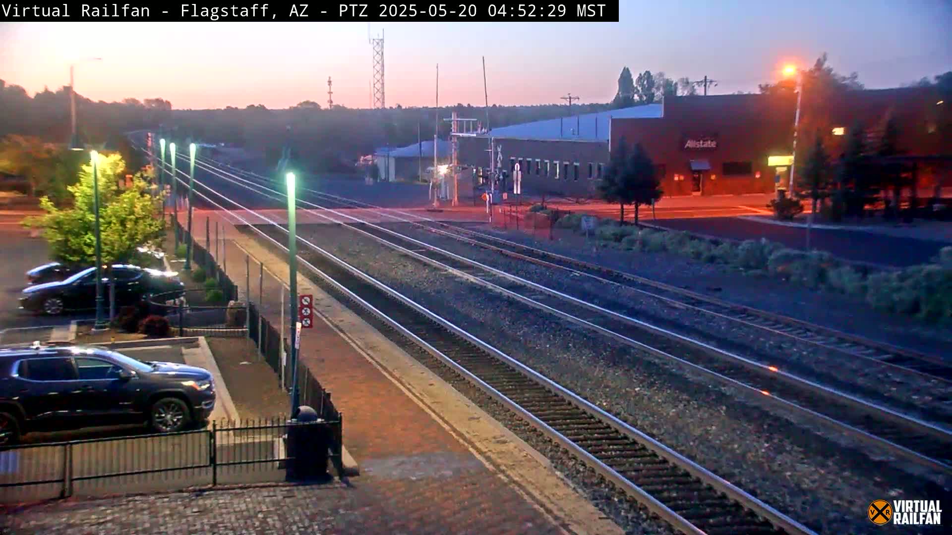 Multiple train tracks extend into the dusky pre-dawn light, past a parking lot with several vehicles and a building.