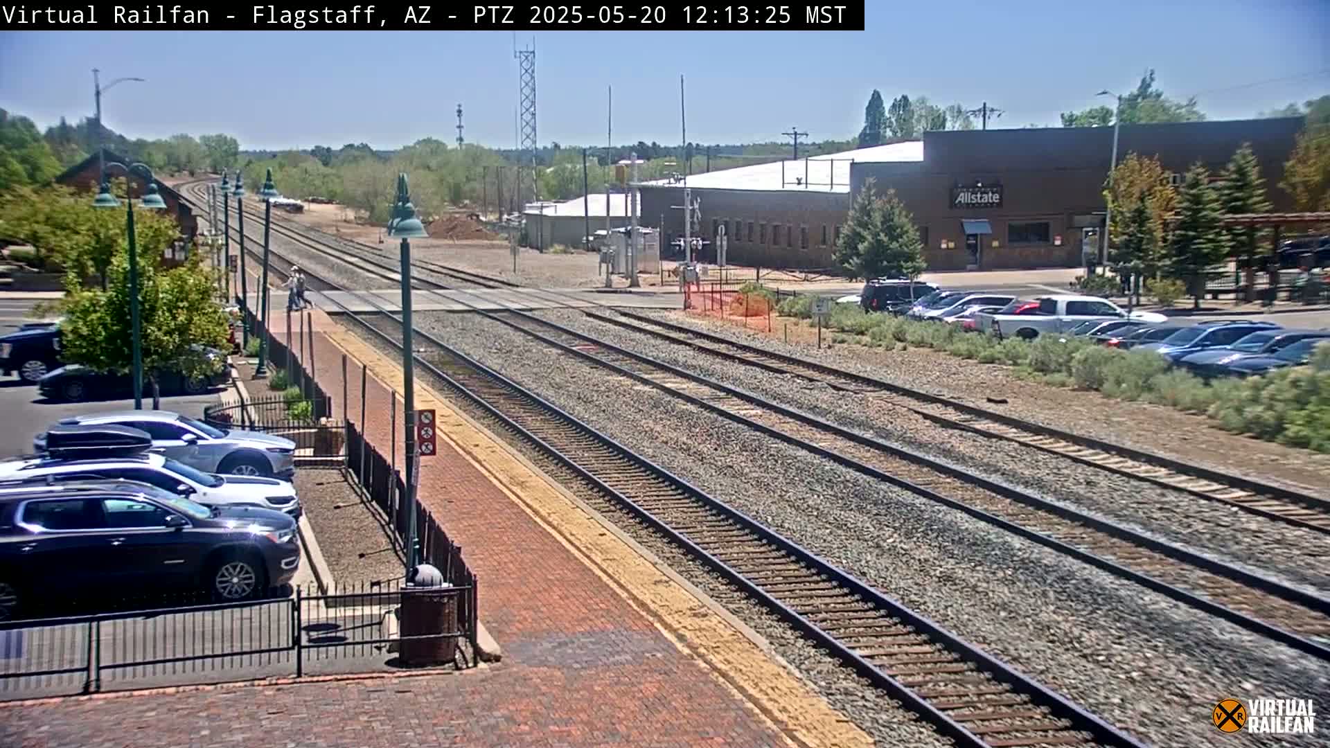A sunny day view of multiple train tracks intersecting with a road, surrounded by cars parked along the side, and a few buildings in the background.