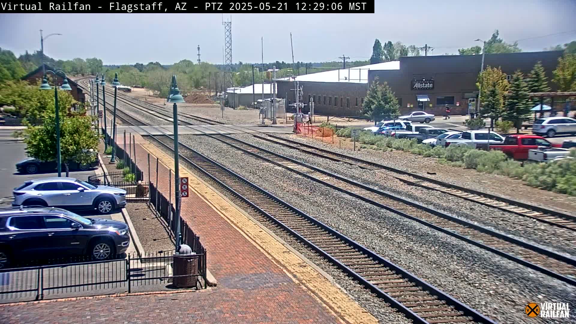 A sunny day shows multiple train tracks extending into the distance, with several cars parked in a lot nearby and a few buildings in the background.