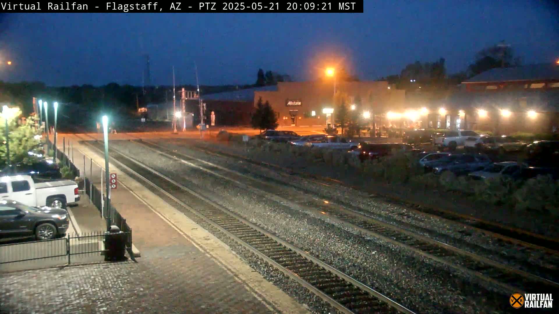 A nighttime view of train tracks at a station, with cars parked in a lot across the tracks and buildings in the background under a dark sky.