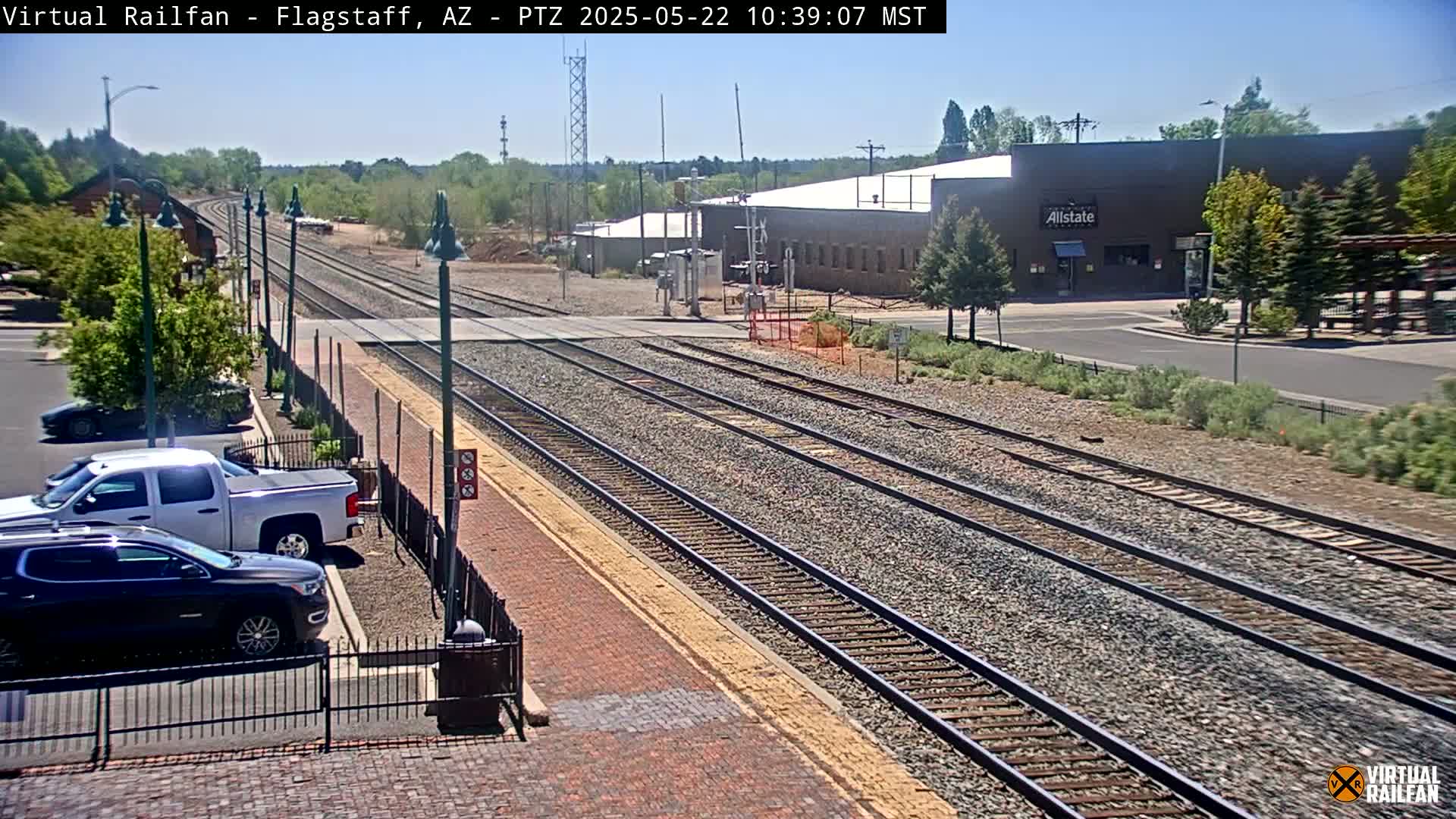 A sunny day view of multiple train tracks extending into the distance, with a parking lot containing several vehicles visible nearby, and a building in the background.