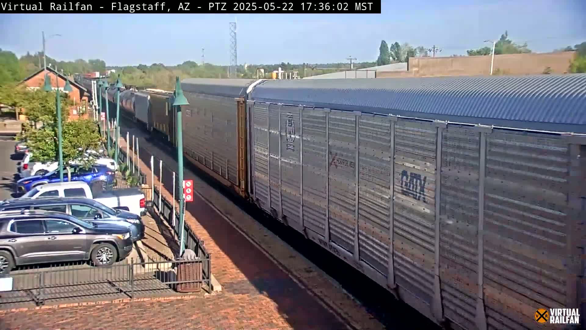 A long freight train passes by a parking lot on a sunny day.