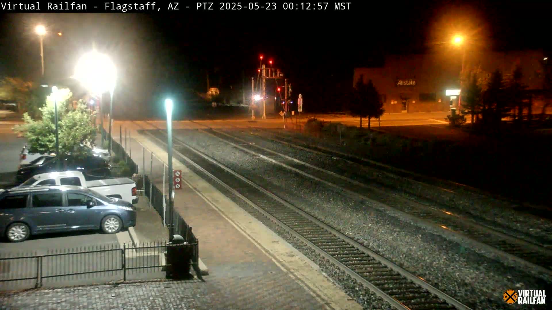 A nighttime view of a train station with several vehicles parked nearby, illuminated by streetlights and under a clear sky.