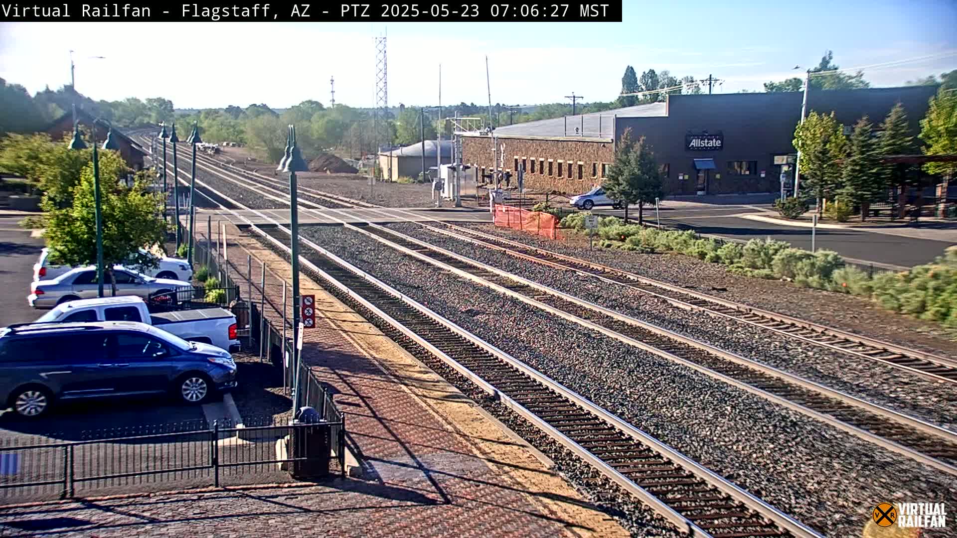 A sunny view of multiple train tracks leading to a building and parking lot with several cars parked nearby.