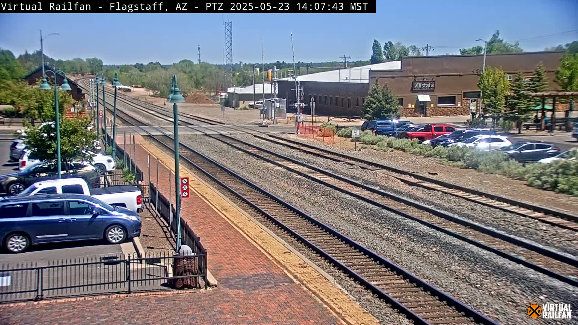 Multiple train tracks extend into the distance under a sunny sky, with parked cars visible alongside a brick roadway and buildings in the background.